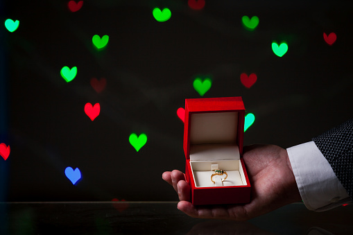 Valentine's Day engagement. A man hand holds a wedding ring in a red box. Behind on a dark background red and green hearts