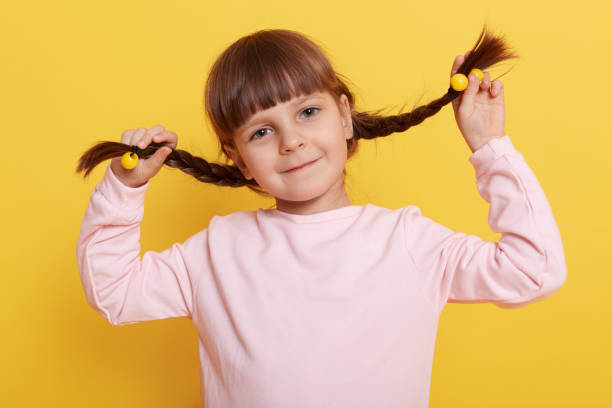 istock Caucasian little girl pulling herself in pigtails while looking directly at camera with cute charming smile, kid in casual pale pink shirt isolated over yellow background. 1298848294