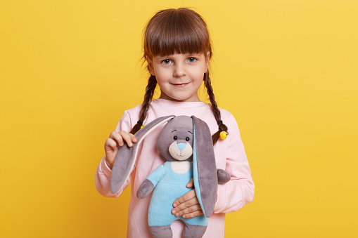 Little kid hugging stuffed toy animal and looks at camera, female kid with fluffy hare against yellow wall, little girl playing with her favorite plaything.