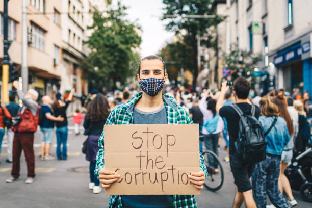 istock Young man with protective face mask, surrounded by a crowd, holding a poster in his hands. Young activist / protester against the corruption. Human rights and social issues concept. 1298849762