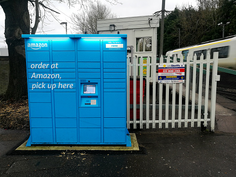 Crawley, UK - 26 January, 2021: color image depicting an Amazon locker goods collection facility outside a train station on a city street in Crawley, UK. A train is speeding past in the background.