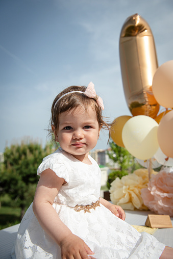Baby is looking at camera surrounded by colored balloons and blue sky.  She is at her birthday party.