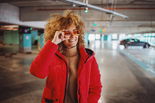 Smiling mixed race hip hop girl in jacket standing in garage and adjusting sunglasses.