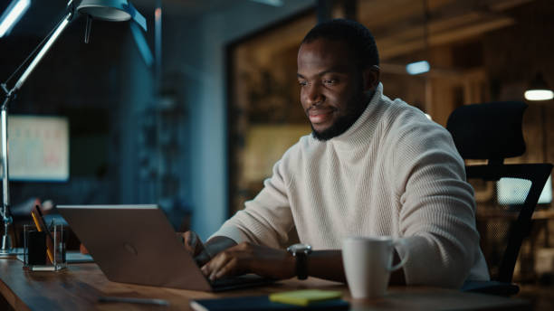 istock Handsome Black African American Man Having an Online Conversation on a Laptop Computer in Creative Office Environment. Happy Male is Browsing Social Media and Replying to Friends in Messenger. 1298861741