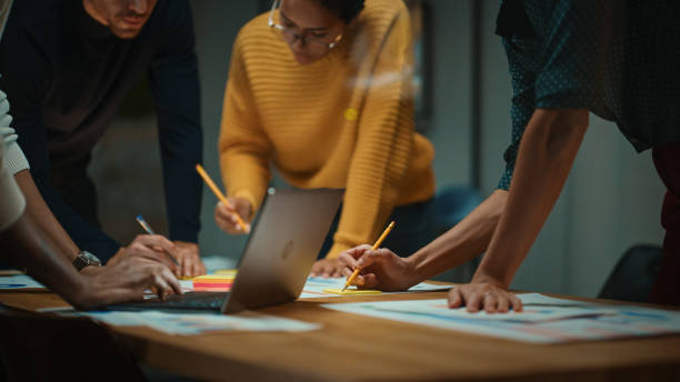 acercarse de un equipo multiétnico diverso que tiene una conversación en la sala de reuniones en una oficina creativa. colegas se apoyan en una mesa de conferencias, mira la computadora portátil y toma notas con lápices en los cuadernos. - mercadotecnia fotografías e imágenes de stock