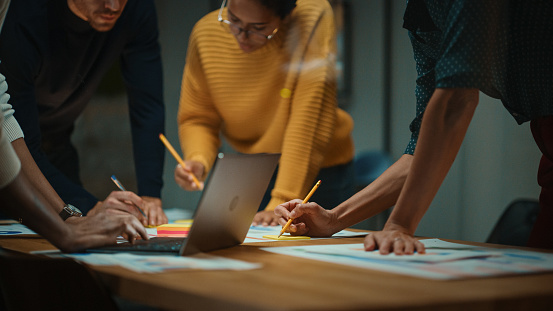 Close Up of Diverse Multiethnic Team Having Conversation in Meeting Room in a Creative Office. Colleagues Lean On a Conference Table, Look at Laptop Computer and Make Notes with Pencils on Notebooks.