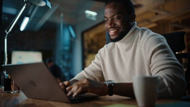 handsome black african american man having an online conversation on a laptop computer in creative office environment. happy male ist browsing social media und antworten auf freunde im messenger. - computer programmer fotos stock-fotos und bilder