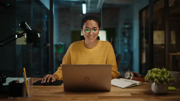 Photo of Portrait of Young Latina Marketing Specialist Working on Laptop Computer in Busy Creative Office Environment. Beautiful Diverse Multiethnic Female Project Manager is Browsing Internet.