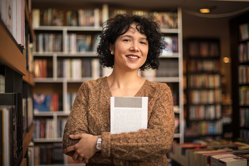 Smiling woman in standing in library and holding book. Looking at camera.