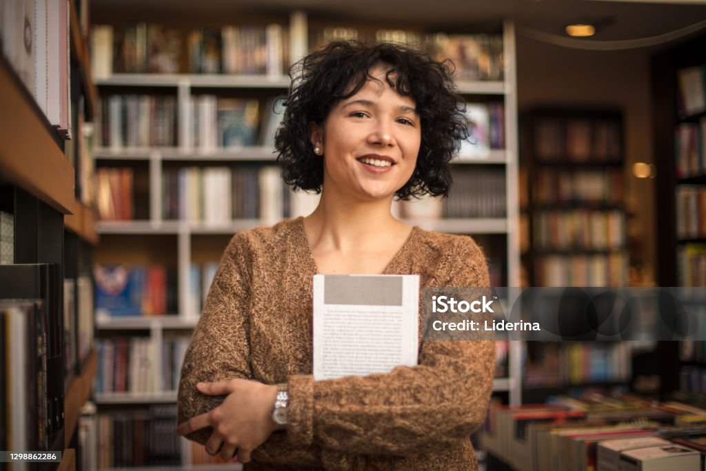 Smiling woman in standing in library and holding book. Looking at camera. Librarian Stock Photo