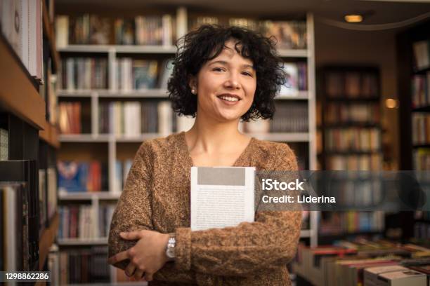 Smiling Woman In Standing In Library And Holding Book Looking At Camera Stock Photo - Download Image Now