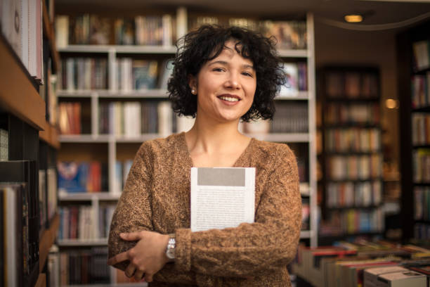 smiling woman in standing in library and holding book. looking at camera. - librarian imagens e fotografias de stock