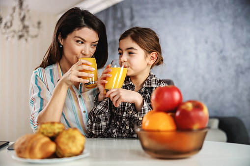 Sonriendo madre e hija sentadas en la mesa del comedor y desayunando saludablemente. Están bebiendo jugo de naranja. photo