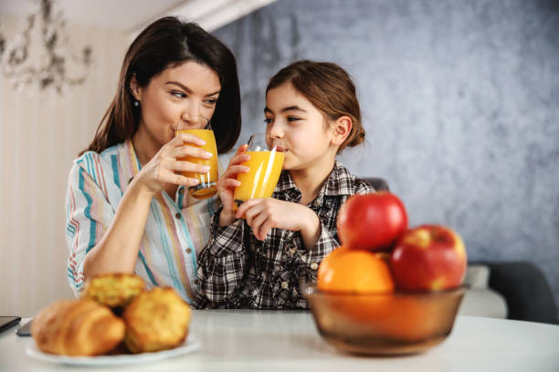 madre e figlia sorridenti sedute al tavolo da pranzo e con una sana colazione. bevono succo d'arancia. - juice glass healthy eating healthy lifestyle foto e immagini stock