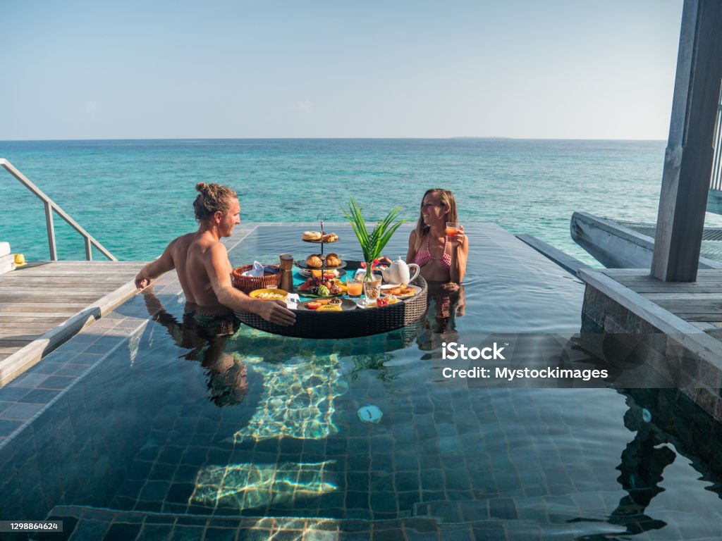 Couple enjoying floating breakfast in private overwater bungalow Couple enjoying tropical vacations from an infinity pool in private over water villa. People travel luxury holidays Maldives Stock Photo