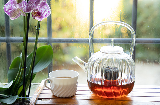A glass teapot with tea and a white cup with tea stand in front of a window