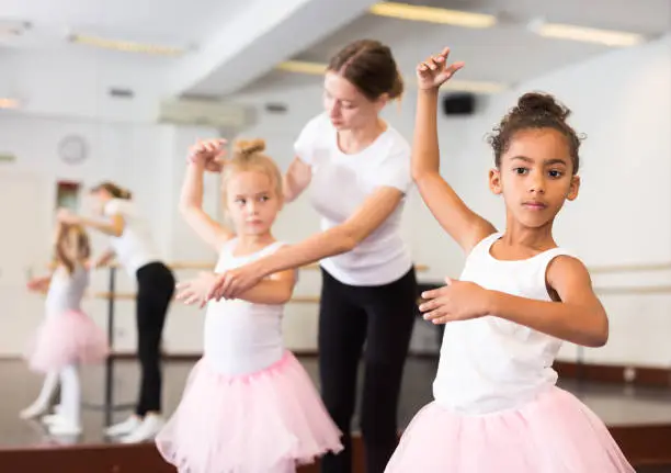 Young female classical dance teacher helping her little girls students in ballet class