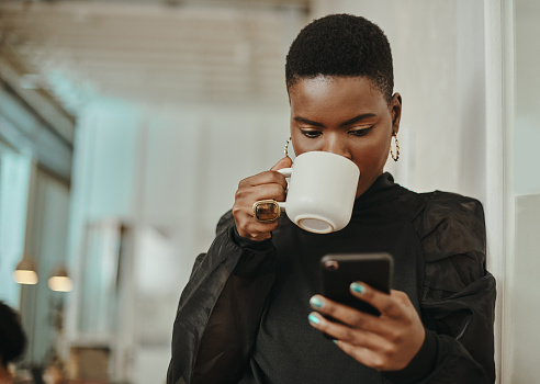 Shot of a young businesswoman having coffee and using a smartphone in a modern office