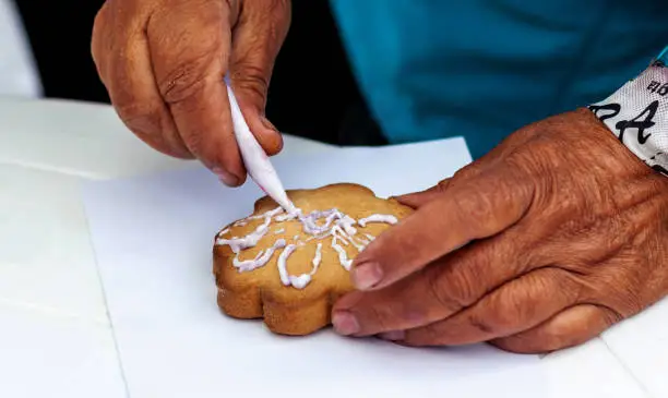 Perm Krai, Russia, July 2019. An elderly woman in a blue jacket with her left hand holds cookies on a white table, with her right hand squeezes white-lilac glaze from a pastry bag