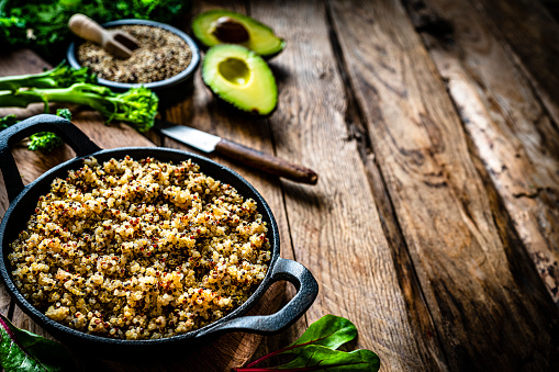 Vegan food: cooked quinoa in a cast iron pan shot on rustic wooden table. Some green vegetables like avocado, broccoli, spinach leaves and lime are around the pan. The composition is at the left of an horizontal frame leaving useful copy space for text and/or logo at the right. Predominant colors are brown and green. High resolution 42Mp studio digital capture taken with SONY A7rII and Zeiss Batis 40mm F2.0 CF lens