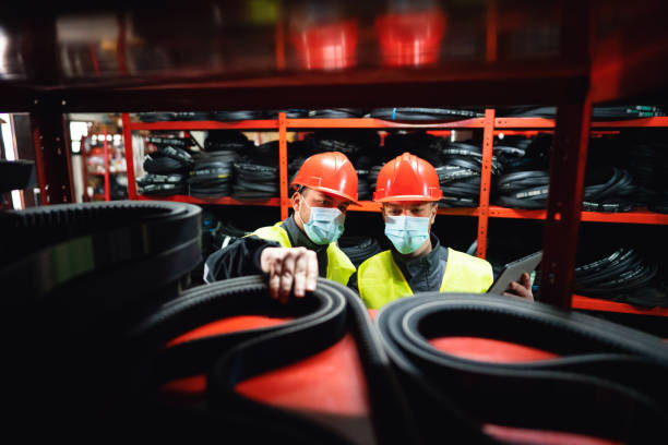 Workes with protective face masks doing stock control by using a digital tablet in a warehouse Two young men, engineers or factory workers, with protective face masks doing stock control by using a digital tablet in a warehouse of endless rubber bands and belts. rubber material stock pictures, royalty-free photos & images