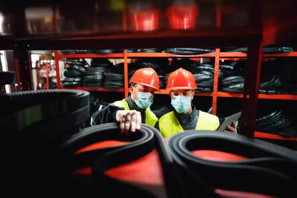 Two young men, engineers or factory workers, with protective face masks doing stock control by using a digital tablet in a warehouse of endless rubber bands and belts.