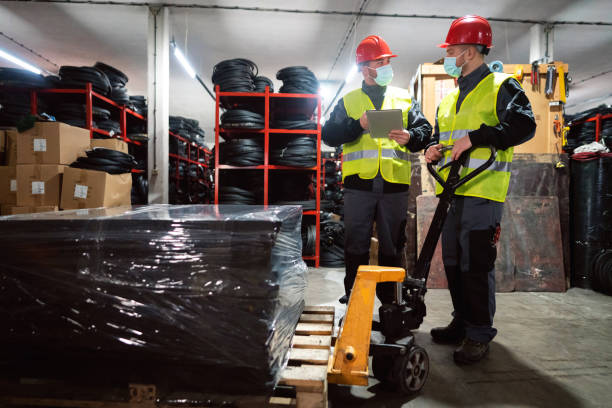 istock Two young male workers with protective face mask sorting finished rubber mats for gym in warehouse by using digital tablet and pallet jack 1298924649