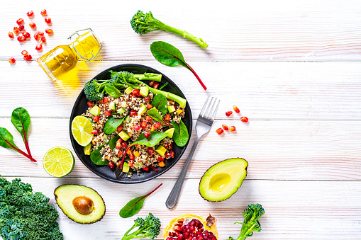 Vegan food backgrounds: healthy fresh multicolored quinoa salad plate shot from above on white table. The salad includes chopped carrot, avocado, pomegranate, broccoli, red chard leaves and tomato. Ingredients are around the plate. The composition is at the left of an horizontal frame leaving useful copy space for text and/or logo at the right. High resolution 42Mp studio digital capture taken with SONY A7rII and Zeiss Batis 40mm F2.0 CF lens