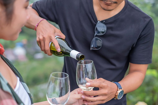 Close up shot of young indonesian man pouring white wine into his glass and the girlfriend glass. Both are standing outside in the nature.