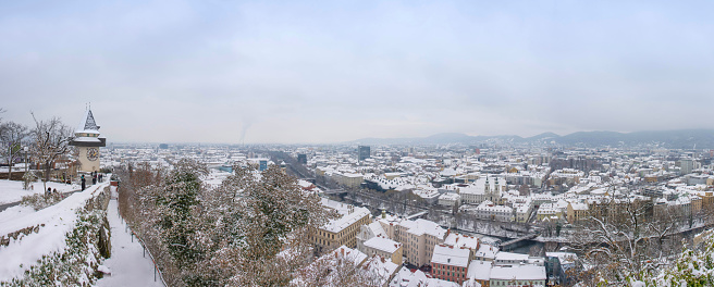 Graz, Austria-December 03, 2020: The famous clock tower on Schlossberg hill and historic buildings rooftops with snow, in Graz, Styria region, Austria, in winter day