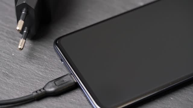 Rotating close-up of charger plugged into turned off smartphone on grey surface.