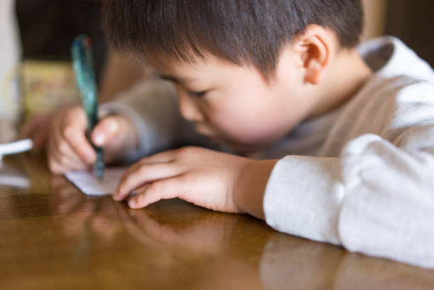Asian boy indoors at home.Writing on a piece of paper with a pen.Writing a letter. Practicing letters.