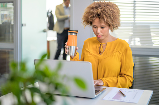 Young businesswoman using laptop while holding water bottle in office.