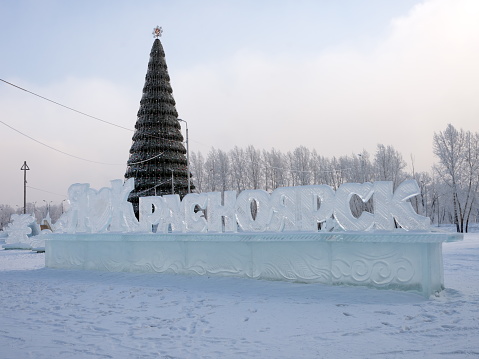 Krasnoyarsk, Krasnoyarsk Territory, RF - January 24, 2018: The name of the city in Russian carved out of ice stands against the background of a Christmas tree decorated with the city's coat of arms.