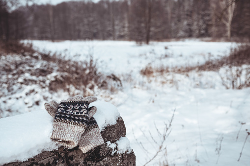 Warm knitted men's gloves lie on a stone barrier covered with snow on a blurred winter background.