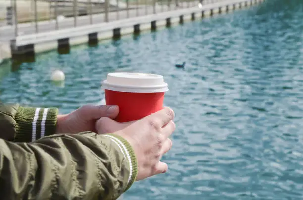 Female hands hold a cardboard cup with coffee on the background of the sea pier. Relaxation by the sea, walks along the coast, takeaway coffee. Space for text.