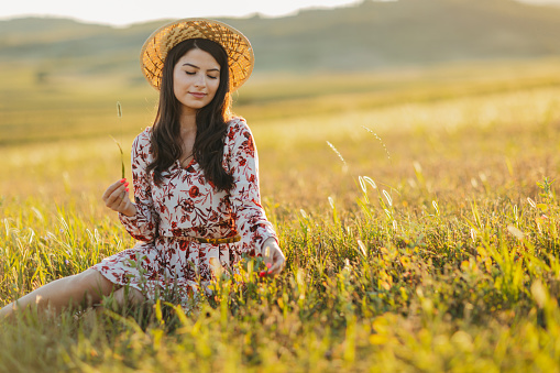 One beautiful young woman in dress with floral pattern and straw hat sitting and enjoying summer sunset over blooming meadow, front view