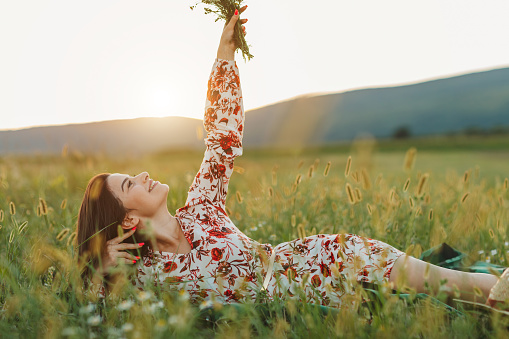 One beautiful young woman with bouquet  of wildflowers, lying on picnic blanket and enjoying summer sunset over blooming meadow, feeling free, relaxed and happy in nature, side view