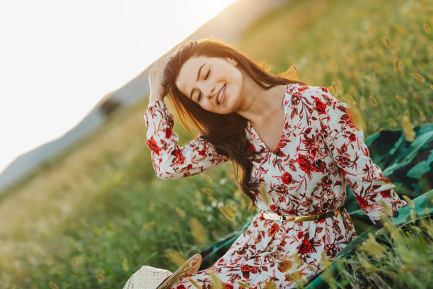 A young woman in a white dress and hat sits on the grass, gently holding a stalk of wild grass while gazing into the distance. The serene setting highlights peace, mindfulness, nature, and quiet reflection
