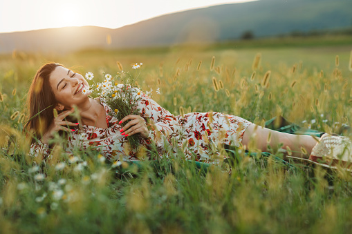 One beautiful young woman with bouquet  of wildflowers, lying on picnic blanket and enjoying summer sunset over blooming meadow, feeling free, relaxed and happy in nature, side view