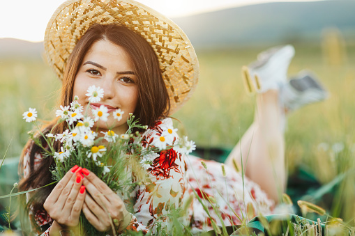 Close-up portrait of one beautiful young woman with sun hat holding and smelling bouquet  of wildflowers, enjoying summer sunset over blooming meadow, feeling free, relaxed and happy in nature, front view