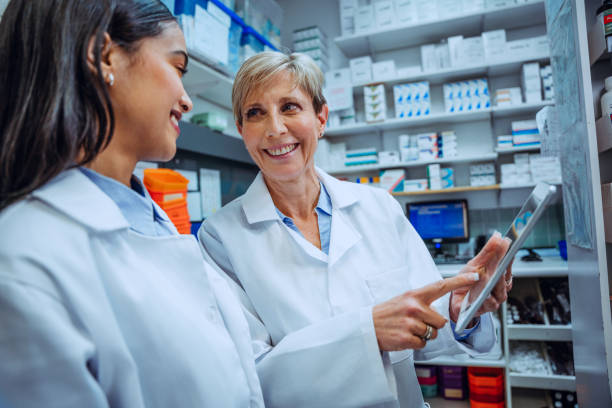 istock Elderly pharmacist teaching new intern student how to work digital tablet standing in medication room in pharmacy 1298970462