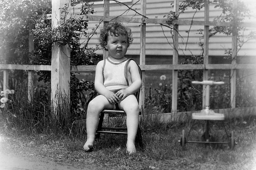 Little girl sitting in chair next to her tricycle in back yard 1928. She is wearing a sunsuit.