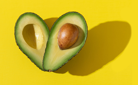 Avocado halves as heart shape close up with hard shadow on yellow background, healthy fats for heart health concept