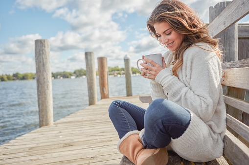 Shot of a beautiful young woman enjoying a warm beverage while relaxing on a bench at a lake