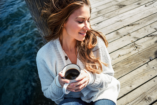 Shot of a beautiful young woman enjoying a warm beverage on a pier at a lake