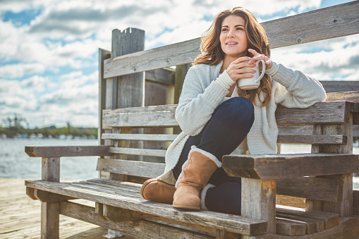 Shot of a beautiful young woman enjoying a warm beverage while relaxing on a bench at a lake