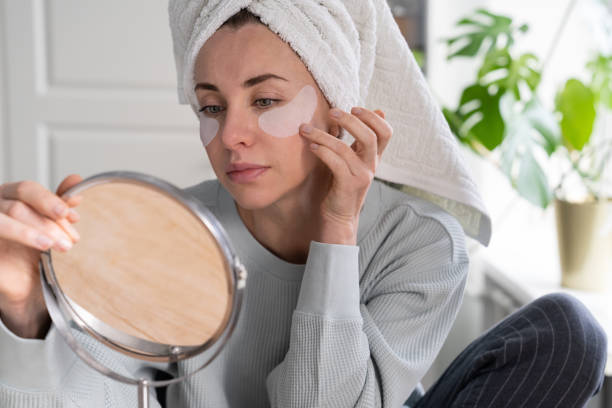 Woman with towel on head apply hydrogel under-eye recovery patches at home, looking in mirror. Woman with towel on head applying patches under eyes enriched with collagen, vitamin E, diminishes the signs of aging, helps reduse eye puffiness, looking in mirror. Face skin care beauty at home. one eyed stock pictures, royalty-free photos & images