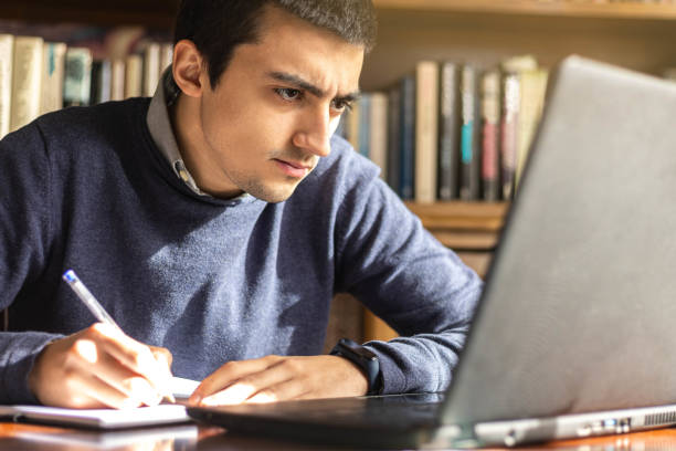 istock Young Caucasian male sitting at a desk working with a laptop taking notes on a webinar or concept training course the computer and studying online 1298990195