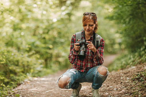 One young woman in plaid shirt crouching in nature and using digital camera on sunny spring day